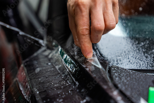 A technician peels clear protective film along a trim line on glossy black bodywork, with water beads and moody shallow depth lighting emphasizing precise craftsmanship.