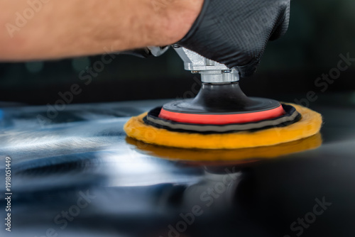 A gloved hand guides a dual action polisher with a yellow pad on a glossy black panel, swirling reflections appear as paint correction progresses under controlled lighting.