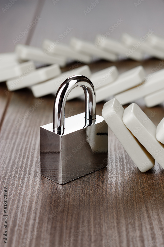 Naklejka premium Standing padlock after domino effect chain reaction. Security concept with lock firmly standing on the brown wooden table with pile of fallen domino tiles in background.
