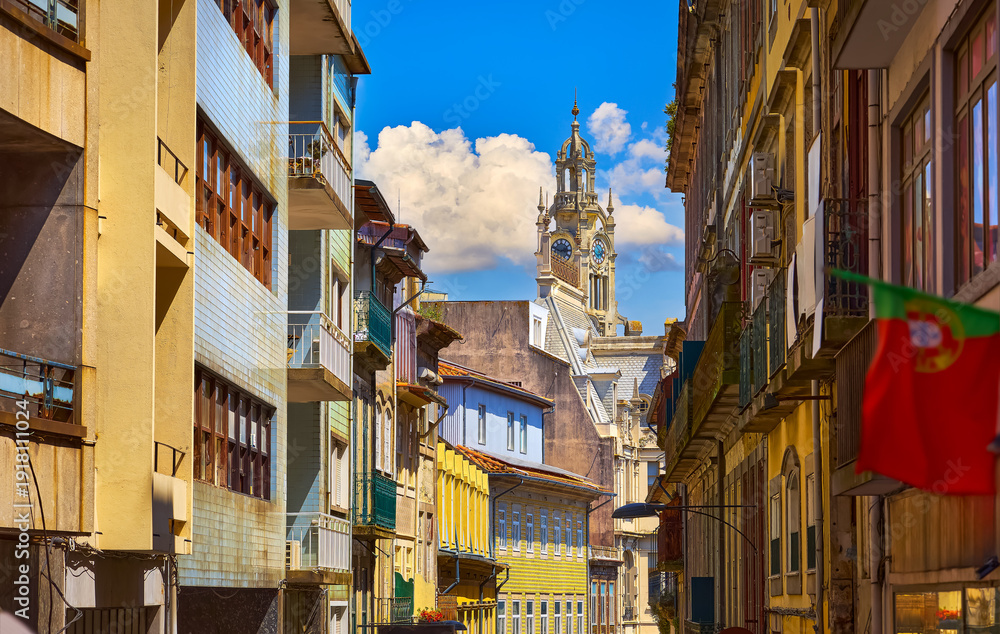 Fototapeta premium Porto, Portugal. Cosy street with traditional colorful houses of old town. View at high medieval tower clock. Sunny summer day. Residential porto city district beautiful antique portugal buildings.