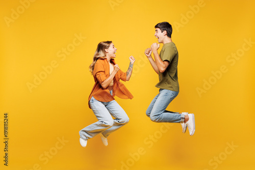 Full body side view young couple friends family man woman wear casual clothes together jump high do winner gesture clench fist isolated on pastel plain light yellow color background studio portrait