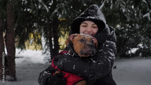 Smiling woman in winter clothes embracing her boxer dog during heavy snowfall in park. Emotional outdoor moment with pet in cold weather.
