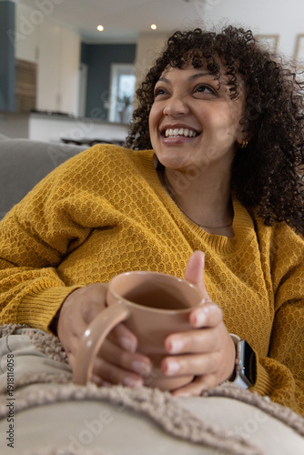 African American woman 20s reclining on gray sofa at home wearing mustard knit holding beige mug