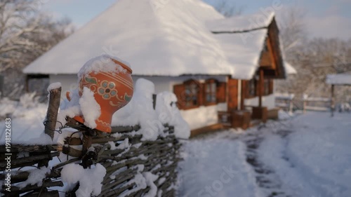 Clay jug covered with snow stands on a wooden fence in winter, while a traditional Ukrainian village house is softly blurred in the background.