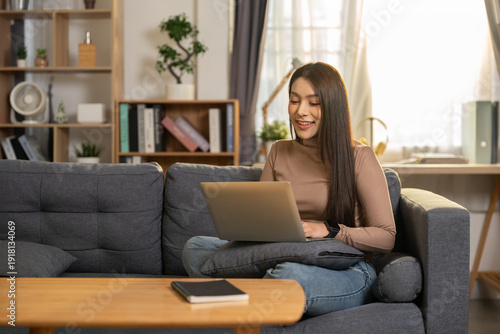 Free Download Happy Asian woman working on laptop sitting on sofa at home. Smiling female freelancer using computer for remote work or online education - GFXLoad