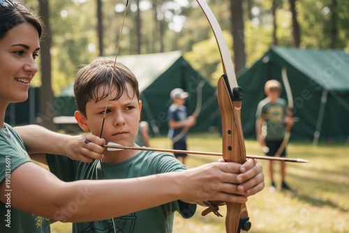 Female instructor teaching young caucasian boy archery at outdoor camp with tents.