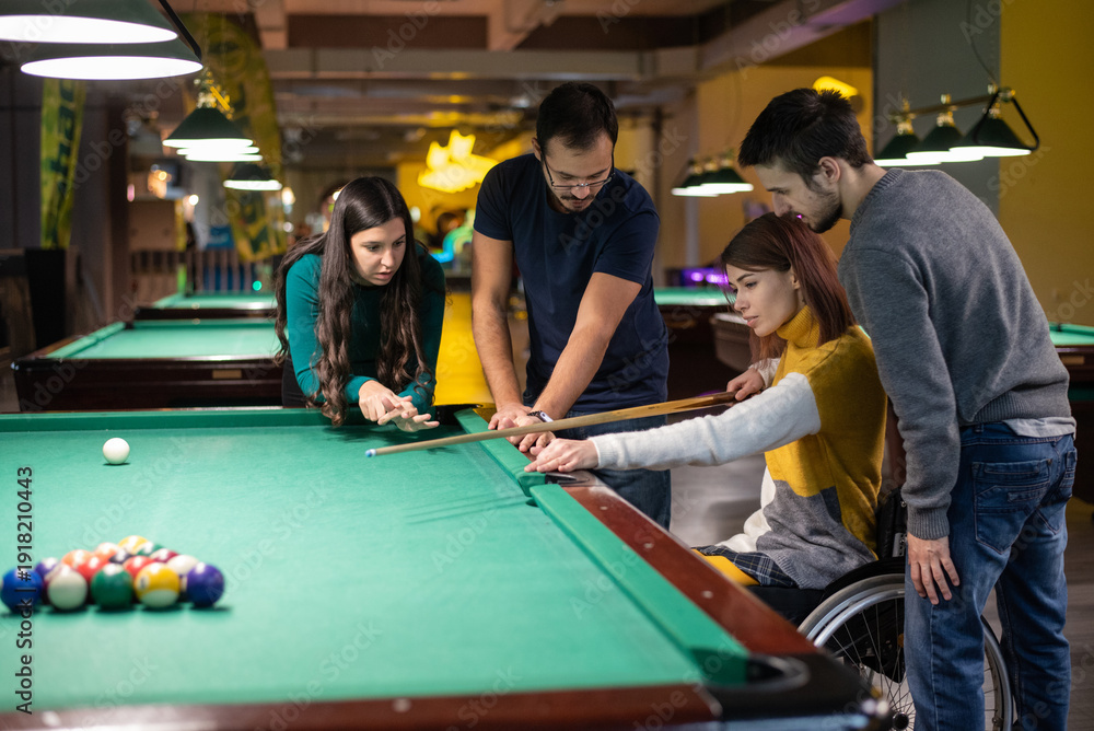 Fototapeta premium Young woman who uses a wheelchair playing billiards with friends