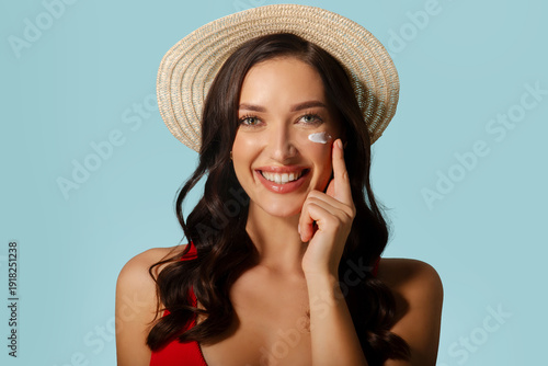 Sun kissed protection. Beautiful woman in straw hat applying cream on her face, smiling at camera, standing against light blue background