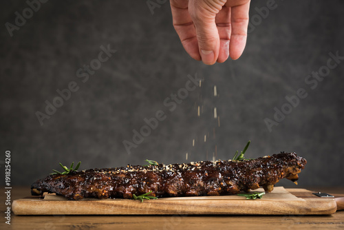 A rack of barbecued spare ribs covered in sauce on a wooden plank with a dark background and a hand dropping extra herbs and seasoning on top of ribs
