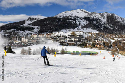 Skiers and snowboarders descending a ski slope in an Italian ski resort. People skiing down a wide snowy piste overlooking Sestriere town in the Piedmont region. Winter holiday resort in the mountains