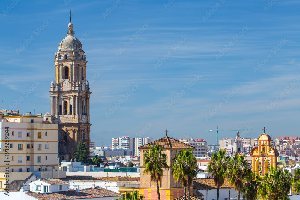 Fototapeta premium Malaga city center skyline with the cathedral tower and yellow church bell tower viewed over rooftops under a clear blue sky with tropical palm trees and urban architecture in Andalusia.