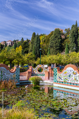 Malaga Spain decorative garden pond in the Pedro Luis Alonso Gardens featuring vibrant ceramic tile art and aquatic plants under a clear blue sky with lush trees.