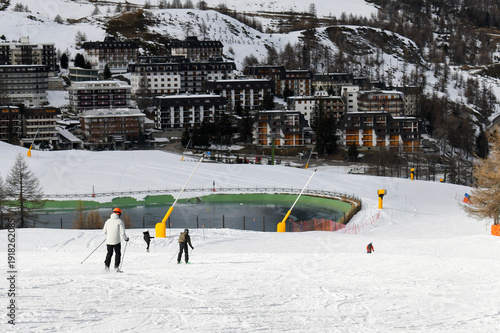 Skiers and snowboarders descending a ski slope in an Italian ski resort. People skiing down a wide snowy piste overlooking Sestriere town in the Piedmont region. Winter holiday resort in the mountains