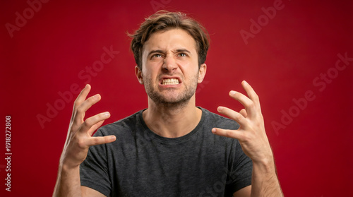 Furious young Caucasian man clenching his teeth and showing aggressive hand gestures on a red background.