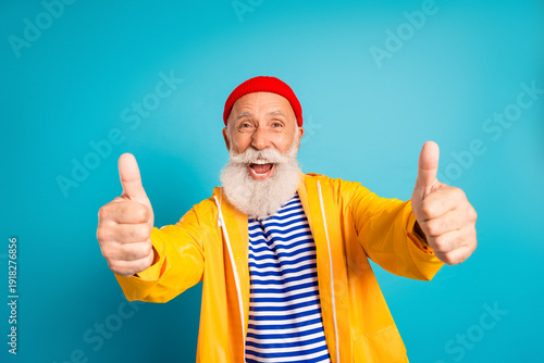 Photos Happy bearded senior man in yellow raincoat gives thumbs up against blue backgro