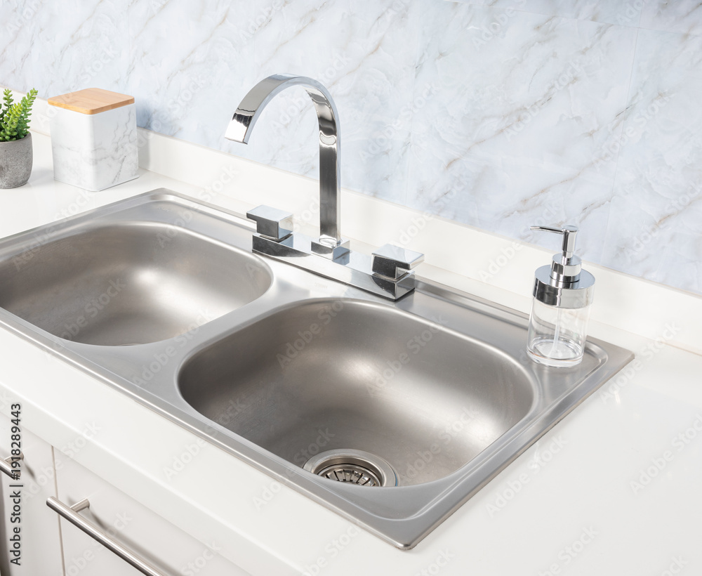 Fototapeta premium Close-Up of Double Basin Drop-In Stainless Steel Kitchen Sink and Faucet Mounted on a White Sink Base Cabinet in Bright Modern Kitchen, White Marble Background.