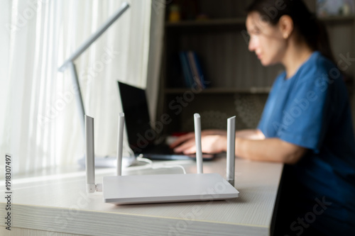 White WiFi Router in Foreground with Woman Working on Laptop in Blurred Background at Home Office