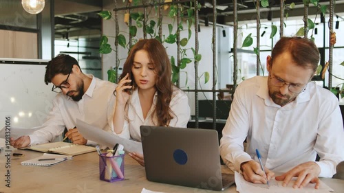 Three coworkers collaborate in an office space. One businesswoman talks on the phone while others write notes and use a laptop to share information.
