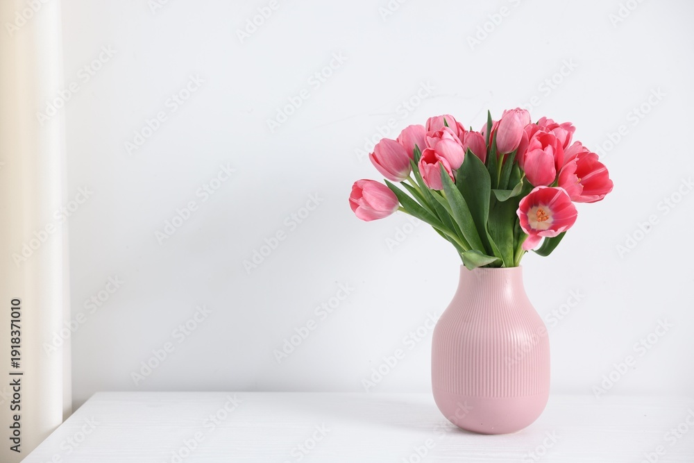 Obraz premium Bouquet of pink tulips in vase on table near white wall. Space for text