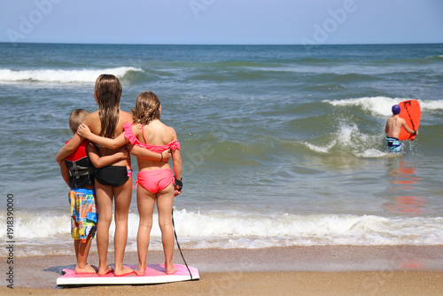Three children embrace, balancing on a boogie board, at the shoreline as waves roll in, capturing carefree summer travel, family connection, the endless joy of beach vacation moments by the ocean.
