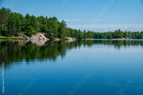 Calm rippled lake water mirrors dense pine trees and a smooth granite shoreline under a bright blue summer sky. Tranquil, pristine wilderness for relaxation and travel cottage themes.