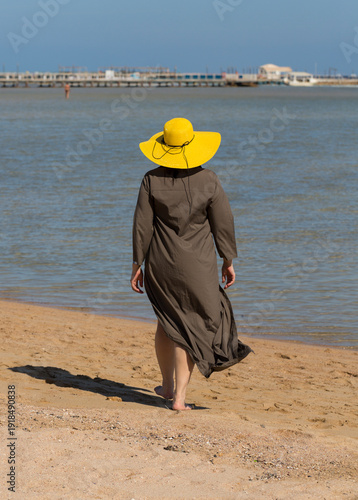 A woman in a long dress and a yellow hat walks along the coastline of the Red Sea. Everyday life of tourists. Hurghada, Governorate - Red Sea, Egypt.