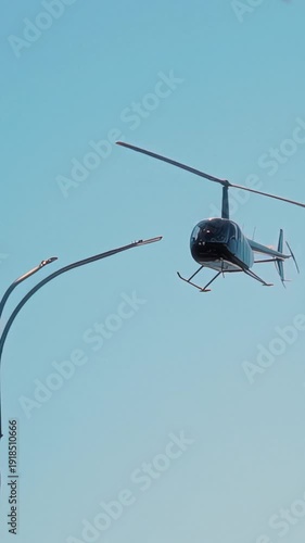 Helicopter flying through clear blue sky, showcasing its rotors in motion, with trees and a streetlight visible in the background, emphasizing aerial movement and flight dynamics