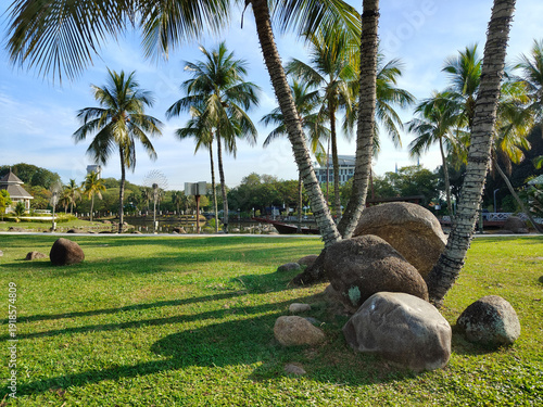 Foto A pile of rocks and coconut trees cast long shadows on the sunny lawn of a tropical garden at  Shah Alam Lake Garden (Taman Tasik Shah Alam) , Selangor, Malaysia