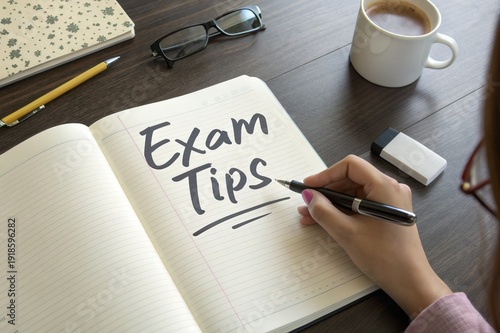 Student writing exam tips in notebook on wooden table with coffee, glasses, and stationery in view