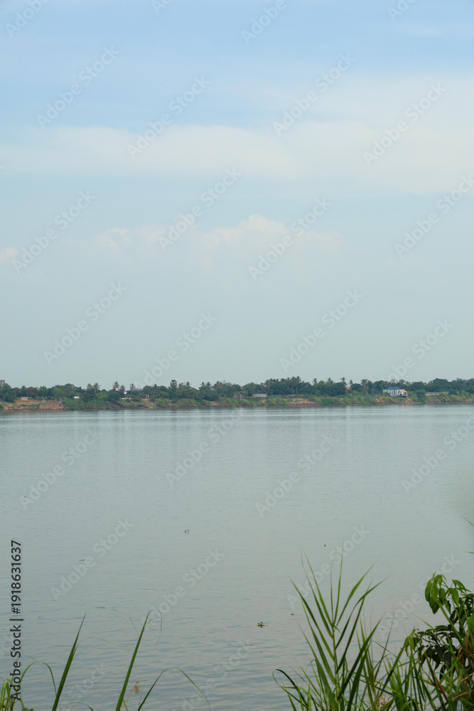Fototapeta premium Serene wide river with calm water reflecting soft blue sky, showcasing distant lush green tropical trees along the quiet bank.