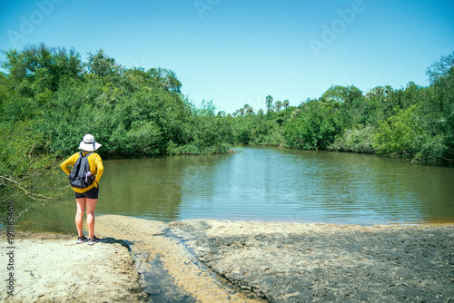 A girl wearing a hat and a yellow shirt is standing in front of a pond in El Palmar National Park