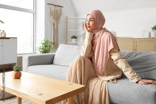 Beautiful young stylish Muslim woman in hijab sitting on sofa at home