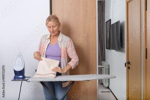 An adult woman folds a freshly ironed pillowcase against a white wall with a wooden panel and a full-length mirror