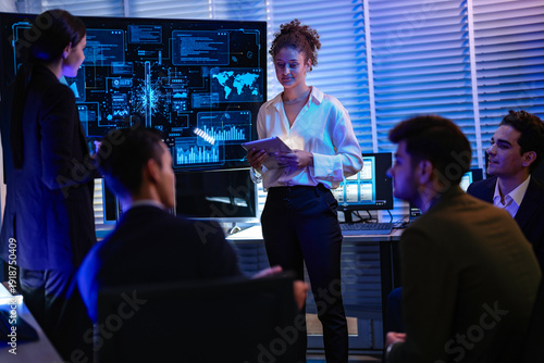 Confident businesswoman presenting global data analysis to team on large digital screen. Professional executive leading IT strategy meeting in modern blue light night office.