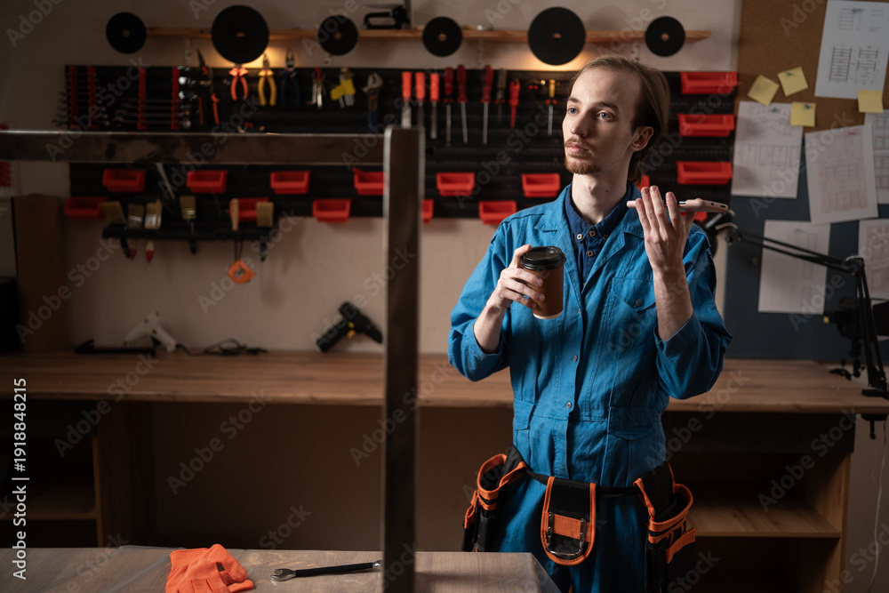Fototapeta premium Handyman in blue overalls resting drinking coffee and communicating with AI assistant in workshop