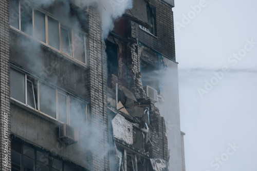 A multi-story residential building exhibiting severe structural damage, broken balconies, and thick smoke following a fire or explosion. High-pressure water is pouring onto the damaged brick façade, a