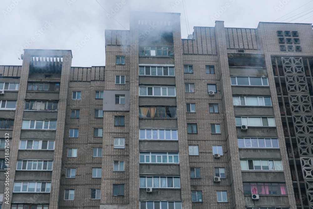 Fototapeta premium A wide shot of a multi-story residential block with smoke rising from the top floors after a major fire. Several windows are broken against a grey winter sky.