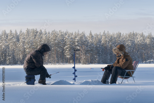 Two fishermen ice fishing on a frozen forest lake in winter. Snow, ice and cold weather create a calm natural outdoor scene. Seasonal winter activity and traditional leisure concept.