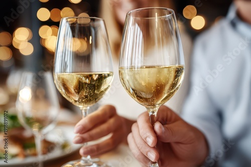 A close-up of two wine glasses filled with white wine, held by a couple at dinner, with warm bokeh lights in the background creating a romantic atmosphere.