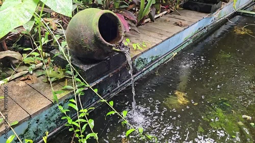 Rustic clay pot water feature pouring into a green pond surrounded by lush plants and wooden planks in a serene garden setting.