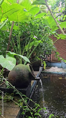 Rustic clay pot water feature in a lush green garden with flowing water and large tropical leaves.