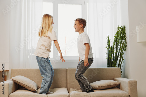 Obraz na plátne Two happy children jumping on a beige sofa in bright living room with natural light