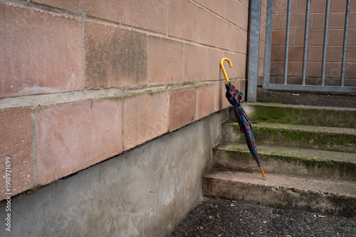 Plaid umbrella on stone stairs leaning against an industrial wall. Everyday urban scene showing a personal accessory, representing mobility, casual style, and outdoor life.