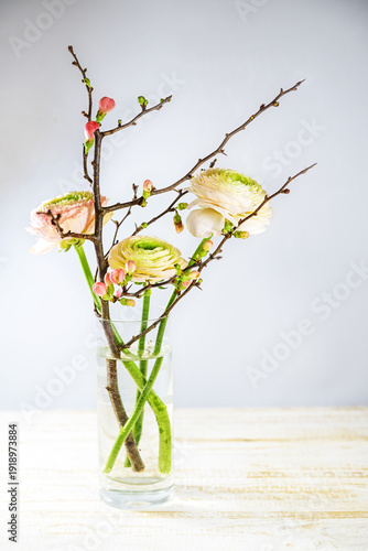 Pastel ranunculus flowers and a quince branch with pink buds in a glass vase against a light gray background, delicate bouquet for spring holidays like Easter, copy space, selected focus, vertical