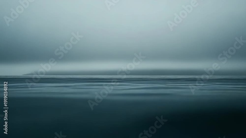 Serene seascape with tranquil water ripples extending to a hazy horizon, while an indistinct landmass emerges from atmospheric mist, evoking feelings of peace and vastness