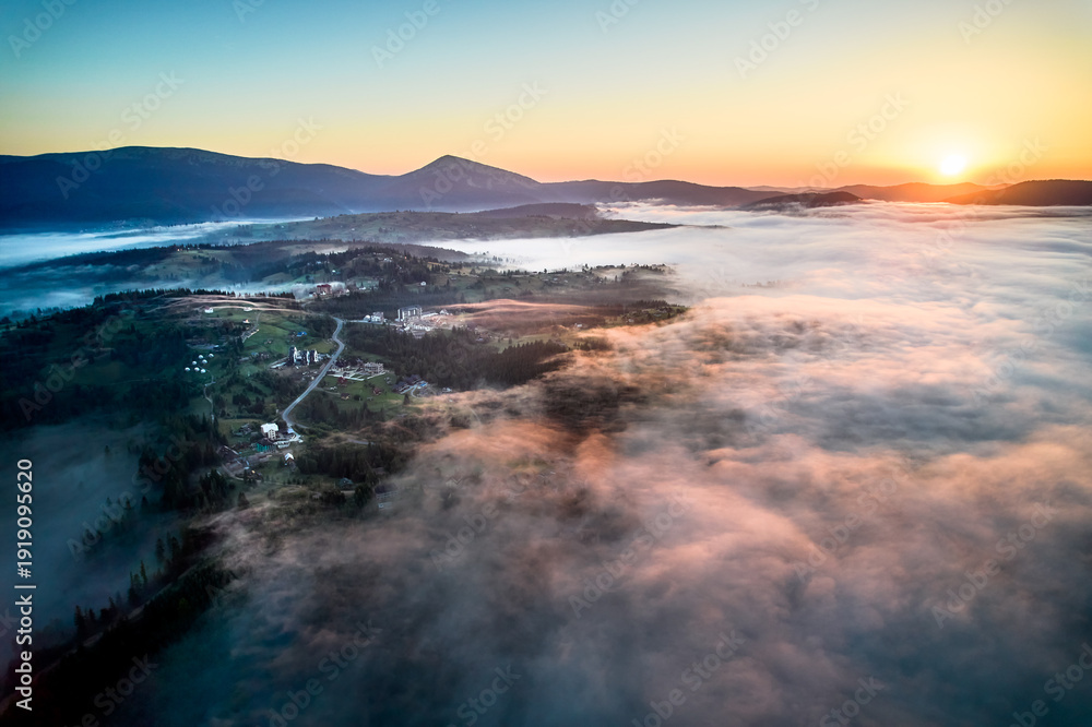 Obraz premium Stunning aerial view of sunrise over mountainous landscape, with thick fog blanketing valleys and hills. Sun's golden rays pierce through mist, casting serene glow over the entire scene.