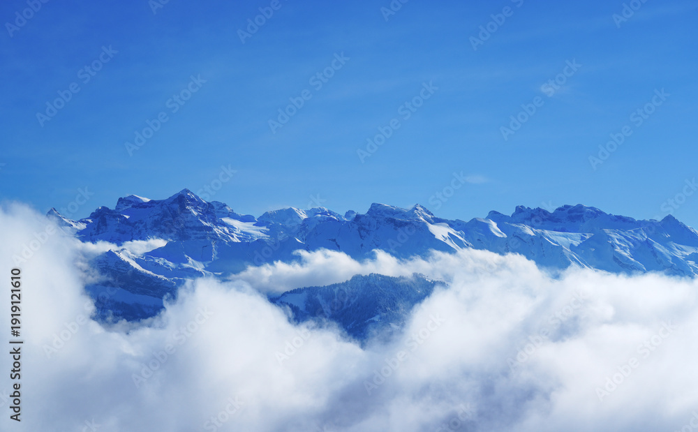 Obraz premium Snow-capped mountain peaks rising above a sea of clouds under a clear blue sky. Temperature inversion creates a dramatic alpine panorama, ideal as a travel or nature background.