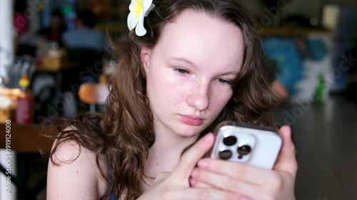 A Slavic European girl with a flower in her hair in the summer with her hair down and a phone in her hands sits thoughtfully and sadly scrolling through her feed.