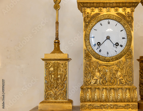 Wallpaper Mural A close-up of an antique gilded mantel clock featuring intricate neoclassical reliefs, winged griffin motifs and a white enamel dial, evoking elegance, history and luxurious handcrafted details Torontodigital.ca