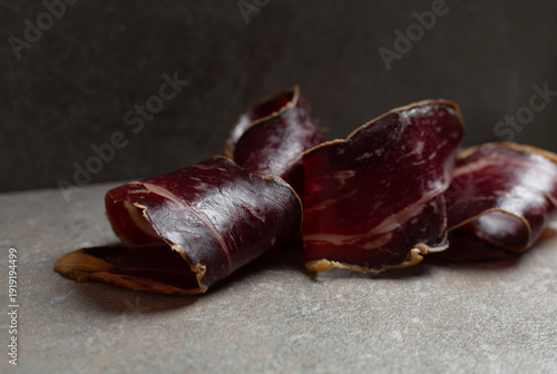 elegant curls of Spanish cured beef cecina presented on a dark stone surface in a sophisticated low-light setting. Premium artisanal charcuterie with rich textures and deep tones